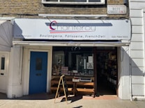 A storefront of a bakery and deli with a white and blue sign reading 'chanteroy' above the entrance, advertising boulangerie, patisserie, and French deli. The store has a glass door, a sign indicating it is closed, and an A-frame chalkboard on the sidewalk. The building exterior is decorated with brick and white painted walls.