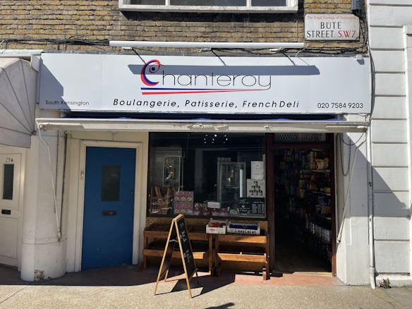 A storefront of a bakery and deli with a white and blue sign reading 'chanteroy' above the entrance, advertising boulangerie, patisserie, and French deli. The store has a glass door, a sign indicating it is closed, and an A-frame chalkboard on the sidewalk. The building exterior is decorated with brick and white painted walls.