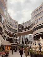 A modern urban scene featuring a striking architectural design with curving glass and steel buildings. Two people are walking in the plaza beneath a skybridge that connects the high-rise structures. The area has outdoor seating with closed umbrellas and some greenery, creating a blend of urban and natural elements.