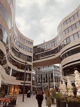 A modern urban scene featuring a striking architectural design with curving glass and steel buildings. Two people are walking in the plaza beneath a skybridge that connects the high-rise structures. The area has outdoor seating with closed umbrellas and some greenery, creating a blend of urban and natural elements.