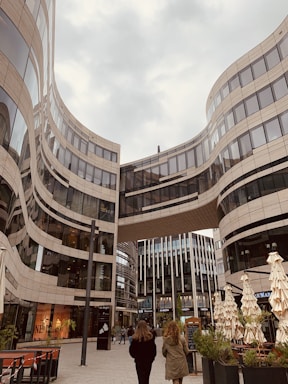 A modern urban scene featuring a striking architectural design with curving glass and steel buildings. Two people are walking in the plaza beneath a skybridge that connects the high-rise structures. The area has outdoor seating with closed umbrellas and some greenery, creating a blend of urban and natural elements.