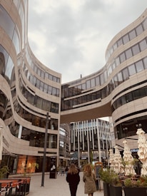 A modern urban scene featuring a striking architectural design with curving glass and steel buildings. Two people are walking in the plaza beneath a skybridge that connects the high-rise structures. The area has outdoor seating with closed umbrellas and some greenery, creating a blend of urban and natural elements.