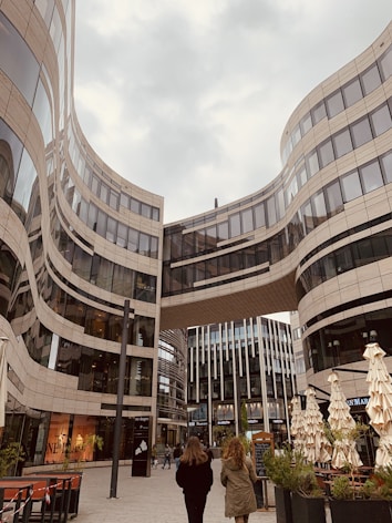 A modern urban scene featuring a striking architectural design with curving glass and steel buildings. Two people are walking in the plaza beneath a skybridge that connects the high-rise structures. The area has outdoor seating with closed umbrellas and some greenery, creating a blend of urban and natural elements.