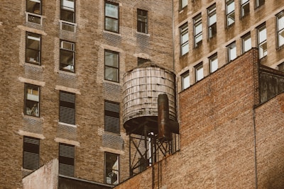 A rustic water tower is perched on the rooftop of an old brick building. The surrounding buildings are tall, with numerous windows, and exhibit a similar brick exterior that showcases an urban architectural style.