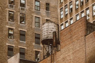 A rustic water tower is perched on the rooftop of an old brick building. The surrounding buildings are tall, with numerous windows, and exhibit a similar brick exterior that showcases an urban architectural style.