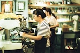 A person wearing a striped shirt and apron is operating an espresso machine in a coffee shop. Another person in similar attire is visible in the background, attending to tasks. Shelves filled with various coffee-related items line the wall.
