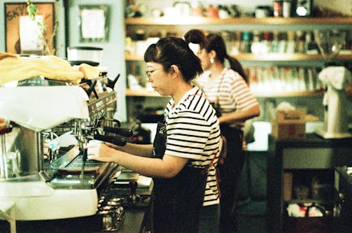 A person wearing a striped shirt and apron is operating an espresso machine in a coffee shop. Another person in similar attire is visible in the background, attending to tasks. Shelves filled with various coffee-related items line the wall.