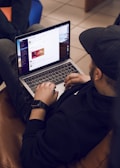 A person is sitting on a brown couch while using a laptop. The individual is wearing a black hoodie and a cap, and appears to be browsing a website or app. The web page shows some sort of promotional or advertisement content. There is another person partially visible, sitting nearby on a blue chair.
