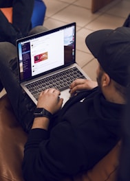 A person is sitting on a brown couch while using a laptop. The individual is wearing a black hoodie and a cap, and appears to be browsing a website or app. The web page shows some sort of promotional or advertisement content. There is another person partially visible, sitting nearby on a blue chair.