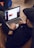 A person is sitting on a brown couch while using a laptop. The individual is wearing a black hoodie and a cap, and appears to be browsing a website or app. The web page shows some sort of promotional or advertisement content. There is another person partially visible, sitting nearby on a blue chair.
