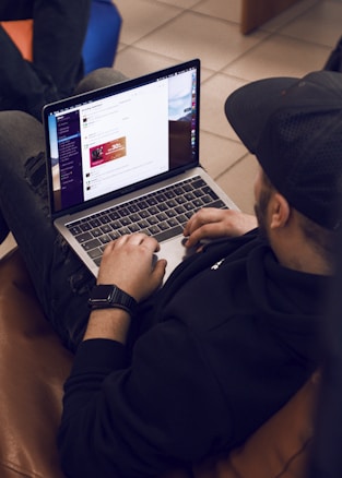A person is sitting on a brown couch while using a laptop. The individual is wearing a black hoodie and a cap, and appears to be browsing a website or app. The web page shows some sort of promotional or advertisement content. There is another person partially visible, sitting nearby on a blue chair.