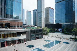 A bustling urban plaza is surrounded by multiple high-rise buildings with glass facades. Many people are walking around the plaza, some in small groups while others are alone. The buildings are modern, featuring a lot of steel and glass. Trees and green areas are visible in the background, adding a touch of nature to the cityscape. There is a retail store with a noticeable logo of a jumping man on the left side of the image.
