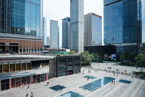 A bustling urban plaza is surrounded by multiple high-rise buildings with glass facades. Many people are walking around the plaza, some in small groups while others are alone. The buildings are modern, featuring a lot of steel and glass. Trees and green areas are visible in the background, adding a touch of nature to the cityscape. There is a retail store with a noticeable logo of a jumping man on the left side of the image.