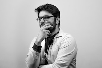 A person wearing glasses and a lab coat appears deep in thought, with a stethoscope around their neck. The image is in black and white.