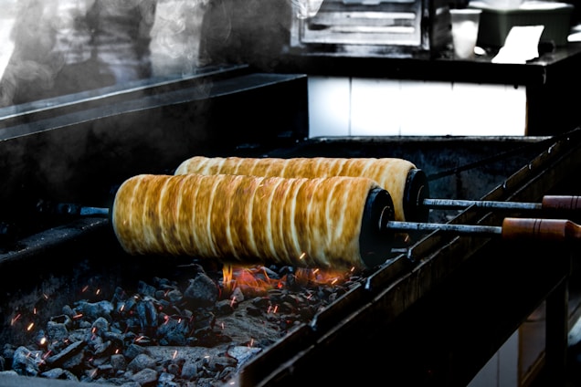 Two large rolls of dough are being cooked over an open charcoal grill. The dough is wrapped around metal skewers, and the charcoal beneath is glowing with red and orange embers. Smoke rises from the grill, creating a rustic atmosphere.
