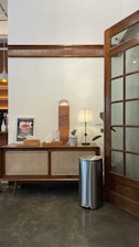 A wooden cabinet topped with a small lamp, a potted plant, a framed artwork, and a skateboard. The setting features a white wall with wooden paneling and a wooden door with glass panes. The floor is polished concrete, and a metallic trash bin is placed beside the cabinet.