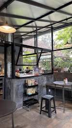 A cozy cafe interior with industrial-style design featuring open glass windows. The setting includes round tables, a black metal stool, and a small cart with coffee-related items. Plants adorn the windowsill, and there is a warm ambiance created by hanging ceiling lights.