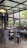 A cozy cafe interior with industrial-style design featuring open glass windows. The setting includes round tables, a black metal stool, and a small cart with coffee-related items. Plants adorn the windowsill, and there is a warm ambiance created by hanging ceiling lights.