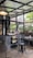 A cozy cafe interior with industrial-style design featuring open glass windows. The setting includes round tables, a black metal stool, and a small cart with coffee-related items. Plants adorn the windowsill, and there is a warm ambiance created by hanging ceiling lights.