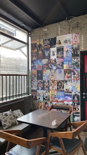 A cozy corner in a cafe with a wooden table and chairs set against a wall covered in a collage of colorful posters, mainly featuring movie and anime themes. Sunlight filters through a large window with metal bars, casting soft shadows inside. Decorative cushions are placed on a bench along the wall, enhancing the inviting atmosphere.