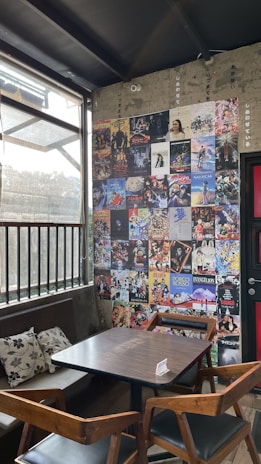 A cozy corner in a cafe with a wooden table and chairs set against a wall covered in a collage of colorful posters, mainly featuring movie and anime themes. Sunlight filters through a large window with metal bars, casting soft shadows inside. Decorative cushions are placed on a bench along the wall, enhancing the inviting atmosphere.