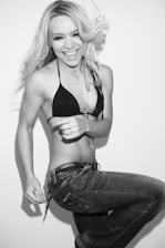 A person with long blonde hair is joyfully laughing. They are wearing a black bikini top and jeans, standing against a plain white background. The person's dynamic pose shows movement, and their expression conveys happiness and excitement.