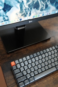 A computer monitor displaying a close-up image of ocean waves sits on a wooden desk. Below the monitor is a mechanical keyboard with grey keys and a single prominent orange key. The keyboard includes keys labeled with letters, numbers, and various symbols.