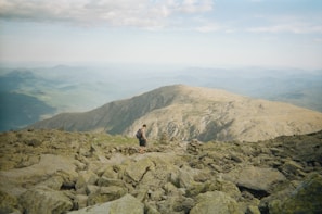 A solitary hiker traverses a rugged rocky trail atop a mountain range, surrounded by expansive views of distant ridges and valleys under a partly cloudy sky.