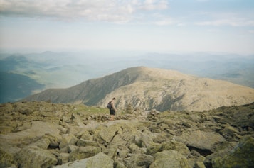 A solitary hiker traverses a rugged rocky trail atop a mountain range, surrounded by expansive views of distant ridges and valleys under a partly cloudy sky.