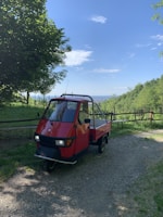 A red three-wheeled vehicle parked on a gravel path surrounded by lush greenery. The background features a clear blue sky with scattered clouds and distant hills visible.