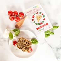 A white dish with a savory cake slice topped with crumbs and surrounded by cherry tomatoes and basil leaves. A box labeled 'Cakes Salés Bio' showing a savory cake picture is placed nearby. Fresh whole garlic, a wooden spoon, and a bunch of ripe vine tomatoes are also visible.