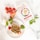 A white dish with a savory cake slice topped with crumbs and surrounded by cherry tomatoes and basil leaves. A box labeled 'Cakes Salés Bio' showing a savory cake picture is placed nearby. Fresh whole garlic, a wooden spoon, and a bunch of ripe vine tomatoes are also visible.
