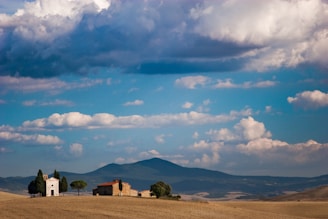 A small chapel and a traditional stone building are situated on a gently rolling hill within a vast landscape. Fluffy clouds populate the bright blue sky, while a mountain range can be seen in the background. A few tall trees surround the buildings, adding to the serene rural setting.