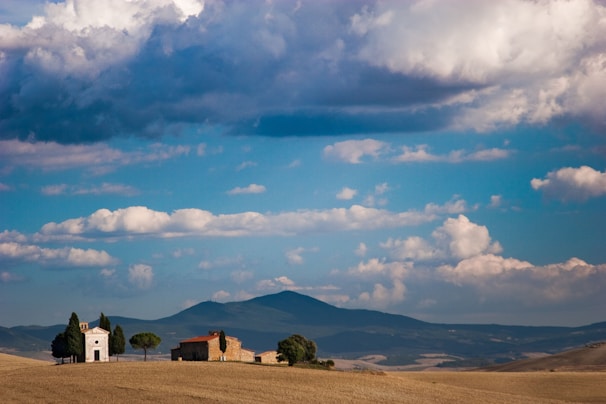 A small chapel and a traditional stone building are situated on a gently rolling hill within a vast landscape. Fluffy clouds populate the bright blue sky, while a mountain range can be seen in the background. A few tall trees surround the buildings, adding to the serene rural setting.