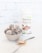A decorative bowl contains coconut-covered chocolate energy balls. Beside the bowl, a spoon holds a small amount of brown protein powder scattered on a white marble surface. A container labeled 'Organic Rice Protein' with 'Nature Zen' branding stands in the background against a subtle white brick wall.