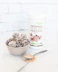 A decorative bowl contains coconut-covered chocolate energy balls. Beside the bowl, a spoon holds a small amount of brown protein powder scattered on a white marble surface. A container labeled 'Organic Rice Protein' with 'Nature Zen' branding stands in the background against a subtle white brick wall.