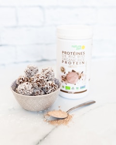 A decorative bowl contains coconut-covered chocolate energy balls. Beside the bowl, a spoon holds a small amount of brown protein powder scattered on a white marble surface. A container labeled 'Organic Rice Protein' with 'Nature Zen' branding stands in the background against a subtle white brick wall.