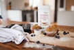 A container of organic pea protein powder sits on a kitchen counter alongside freshly baked muffins and scattered blueberries. The scene is set on a wooden cutting board, surrounded by a striped kitchen towel and a muffin tray. The overall setting suggests a cozy home kitchen environment.
