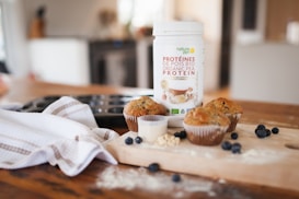 A container of organic pea protein powder sits on a kitchen counter alongside freshly baked muffins and scattered blueberries. The scene is set on a wooden cutting board, surrounded by a striped kitchen towel and a muffin tray. The overall setting suggests a cozy home kitchen environment.