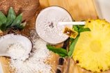 A tropical smoothie adorned with shredded coconut and a slice of pineapple on the rim, surrounded by halved coconuts, grated coconut, a slice of pineapple, and mint leaves on a wooden board.