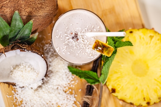 A tropical smoothie adorned with shredded coconut and a slice of pineapple on the rim, surrounded by halved coconuts, grated coconut, a slice of pineapple, and mint leaves on a wooden board.