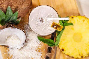 A tropical smoothie adorned with shredded coconut and a slice of pineapple on the rim, surrounded by halved coconuts, grated coconut, a slice of pineapple, and mint leaves on a wooden board.