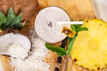 A tropical smoothie adorned with shredded coconut and a slice of pineapple on the rim, surrounded by halved coconuts, grated coconut, a slice of pineapple, and mint leaves on a wooden board.