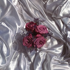 A bouquet of deep pink roses with small purple flowers is arranged on an elegant, glossy silver satin fabric. The soft folds and sheen of the fabric create a luxurious and sophisticated backdrop, highlighting the vibrant colors of the flowers.