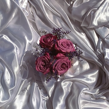 A bouquet of deep pink roses with small purple flowers is arranged on an elegant, glossy silver satin fabric. The soft folds and sheen of the fabric create a luxurious and sophisticated backdrop, highlighting the vibrant colors of the flowers.