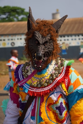 A person wearing a wolf costume with intricate, colorful patterns and detailing, including a lifelike wolf mask with snarling features. The costume is adorned with a variety of colors such as red, yellow, blue, and purple, with a decorative collar featuring small bells. The background shows an outdoor setting with blurred elements and a building facade.