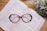A pair of tortoiseshell eyeglasses is placed on top of an open book with visible text on a wooden surface. To the side, a pot of small white flowers with green leaves adds a touch of nature to the scene.