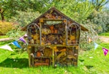 A wooden insect hotel stands in a lush green garden, filled with a variety of natural materials like logs, sticks, pine cones, and clay pots. It has a pitched roof and is surrounded by colorful triangular bunting, creating a festive atmosphere. The structure is set against a backdrop of dense greenery under a bright blue sky.