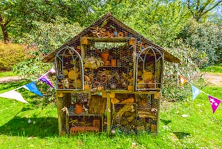 A wooden insect hotel stands in a lush green garden, filled with a variety of natural materials like logs, sticks, pine cones, and clay pots. It has a pitched roof and is surrounded by colorful triangular bunting, creating a festive atmosphere. The structure is set against a backdrop of dense greenery under a bright blue sky.