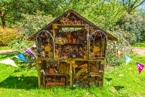 A wooden insect hotel stands in a lush green garden, filled with a variety of natural materials like logs, sticks, pine cones, and clay pots. It has a pitched roof and is surrounded by colorful triangular bunting, creating a festive atmosphere. The structure is set against a backdrop of dense greenery under a bright blue sky.
